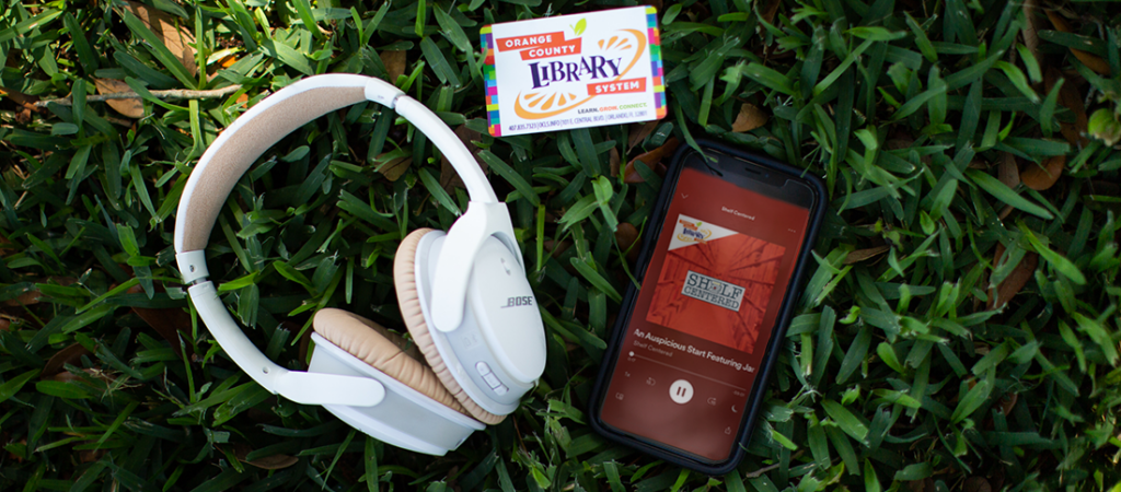 Headphones, library card, and phone with Shelf Centered podcast displayed; on grassy field