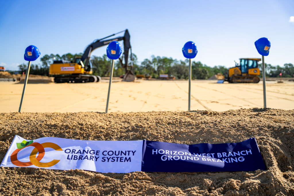 Four golden shovels with blue hard hats on top stand in a pile of dirt in front of construction equipment. A banner with the Orange County Library System Logo and Horizon West Groundbreaking lays on top of the dirt pile.