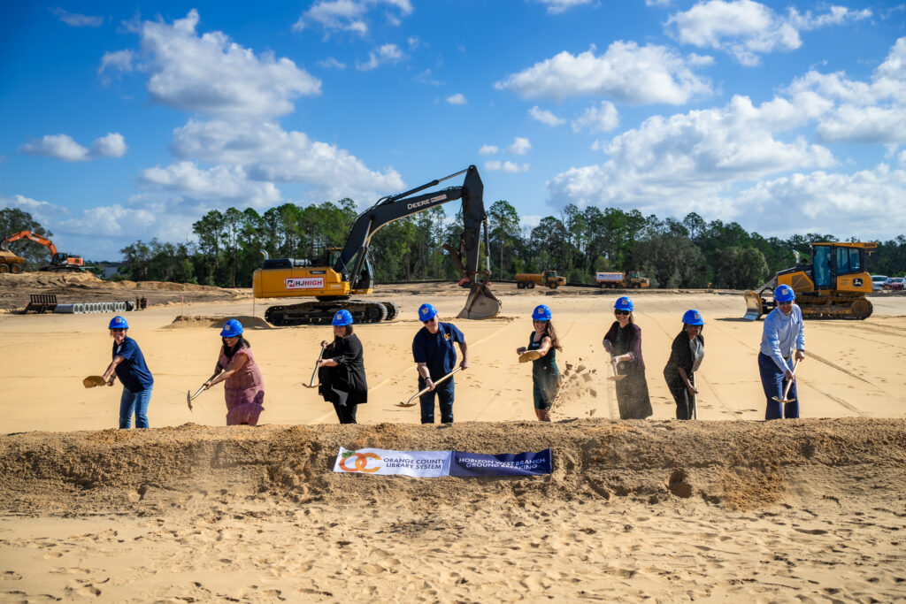 Members of the OCLS Board of Trustees, CEO Steve Powell, and Chief of Neighborhood Services Danielle King wear blue hard hats and throw shovels of dirt in front of construction equipment under a bright blue sky