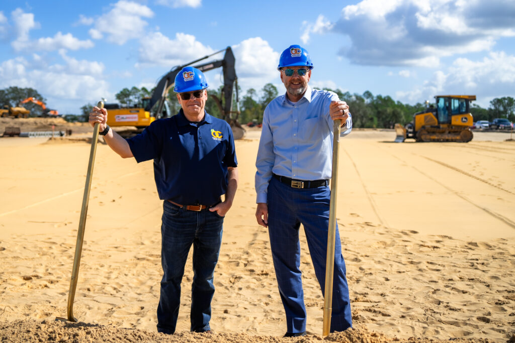 Orange County Library System CEO Steve Powell and Orange County Library System Board of Trustees President Crockett Bohannon wear blue hard hats and hold golden shovels at cleared site in front of construction equipment