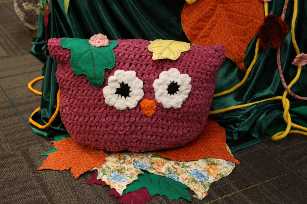 large owl crocheted out of pink yarn with white eyes sitting on fabric leaves