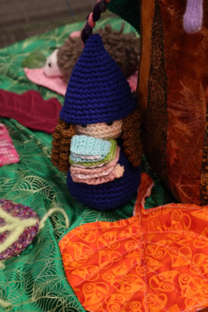 crochet gnome with curly brown hair carrying a pile of books