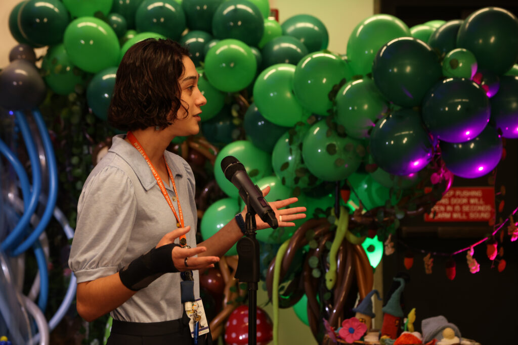 woman with short dark hair standing at a microphone in front of a balloon sculpture