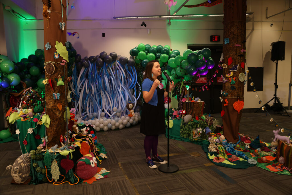 Woman with long dark hair standing at a microphone in front of a fabric and balloon art installation