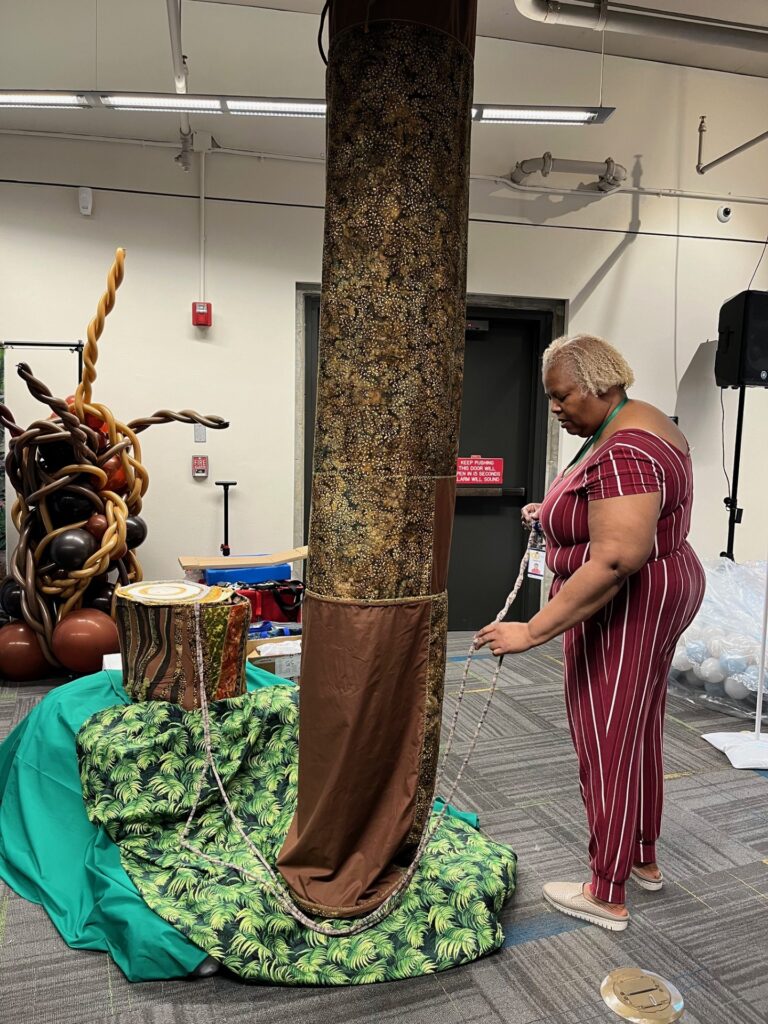 Woman standing next to a fabric tree, soft sculpture tree stump and cloth draped to resemble a forest floor