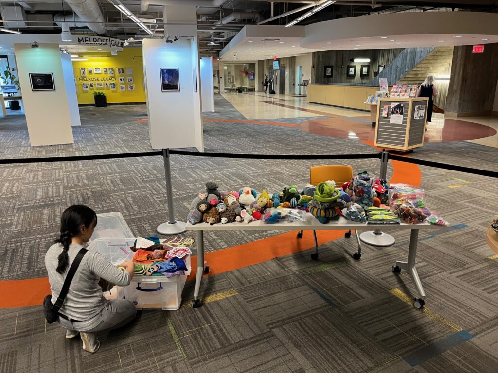 Woman kneeling next to a plastic bin and table full of soft sculpture and fiber arts projects that fit a forest theme