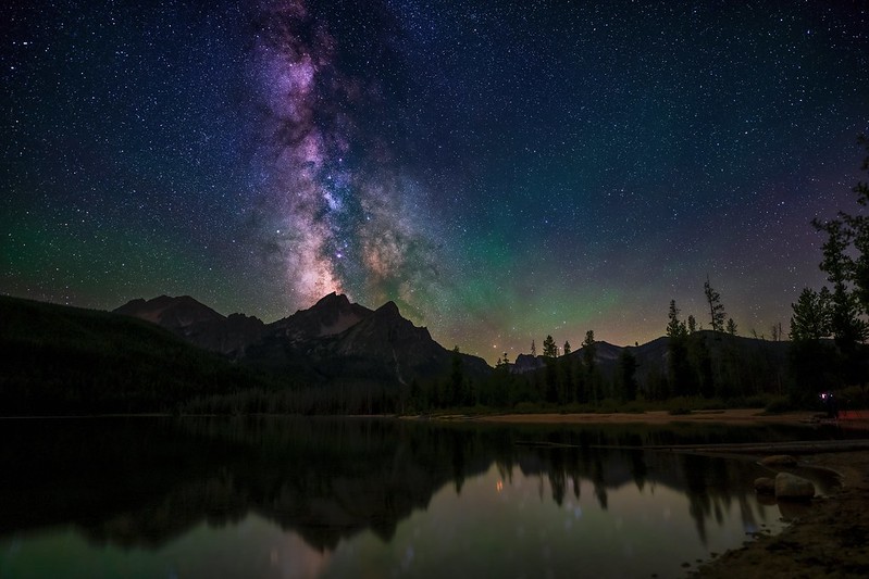 View of starry night sky from across a body of water. Mountains and trees are visible in front of the star-lit sky.