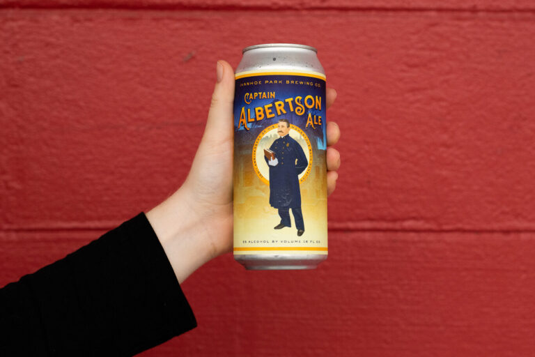 Hand holding a can of Albertson Ale in front of a red wall.