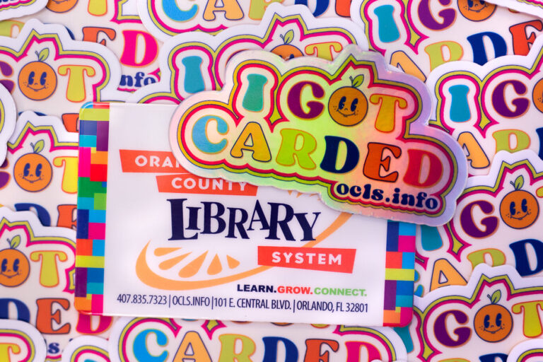 An OCLS library card and ''I Got Carded'' pin sitting on a collage of ''I Got Carded'' stickers.