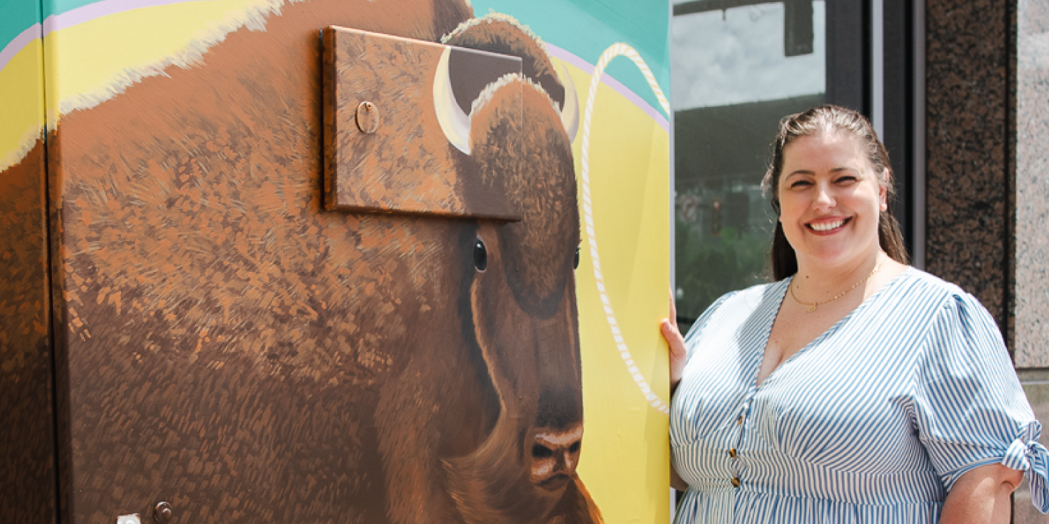 Library graphic specialist Kelly Williams-Cramer standing next to an electrical box featuring her painting of Barney the Buffalo.