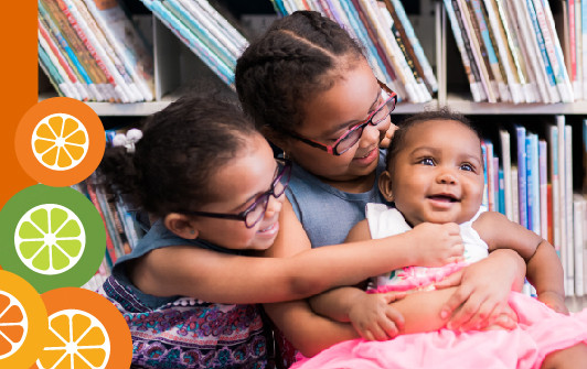 Two young girls holding their baby sister in front of a book shelf.