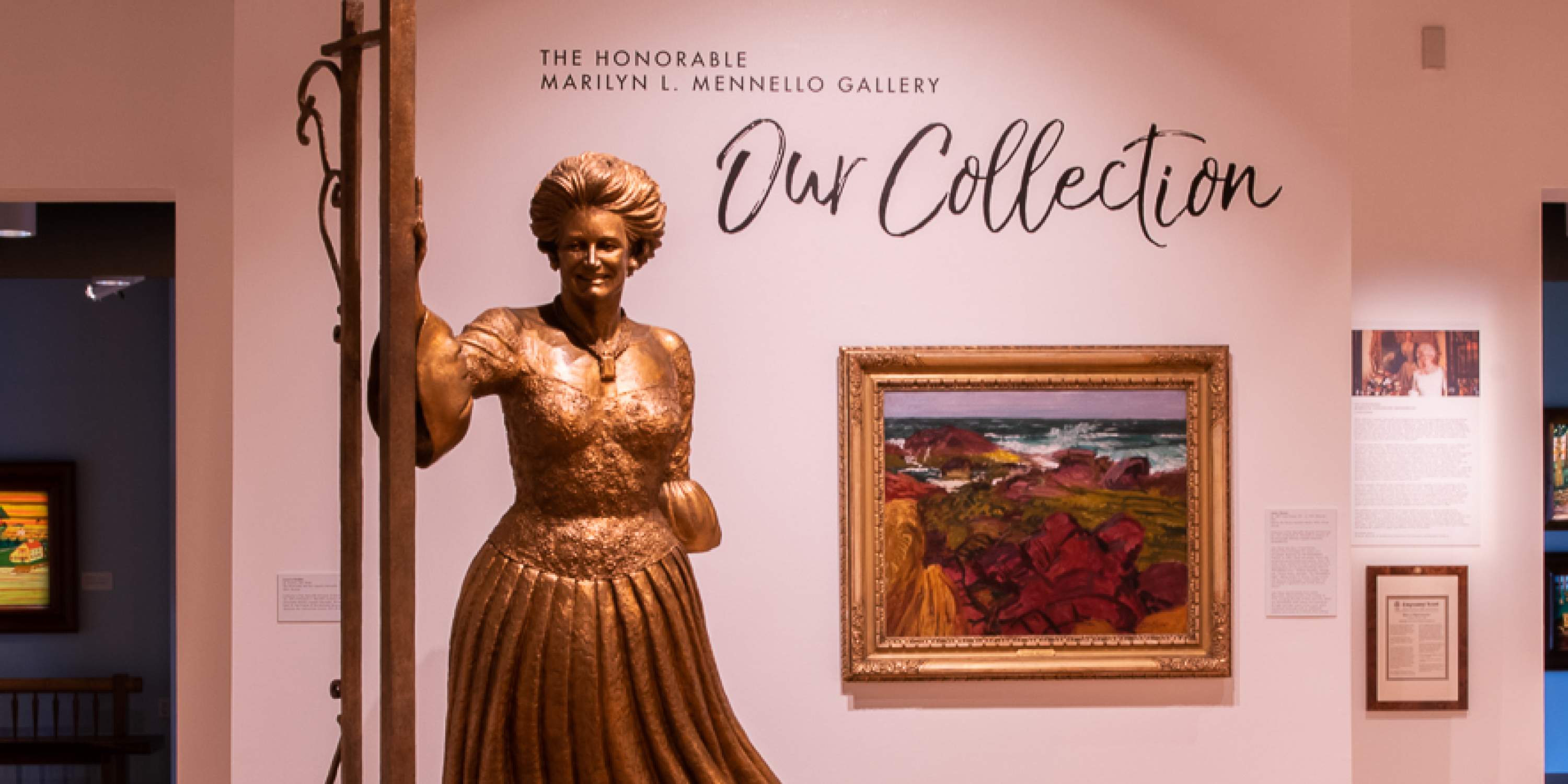 A photograph of the interior of the Mennello Musuem of American Art's Honorable Marilyn L. Mennello Gallery. Bronze statue of Marilyn L. Mennello standing in front of a white wall with mounted paintings.