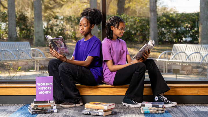 Two young girls sit in front of a large window reading books by women authors for Women's History Month.