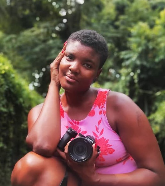 Photograph of Erica Lanier, Photographer. Black woman with short hair wearing a pink sleeveless top, holding a camera.