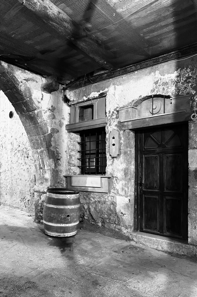 Black and white photograph of an old stone wall, with barred window, antique door, and water barrel. Photo credit: JJ Dennis.