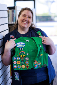 Woman standing next to bookshelf holds green Girl Scout Vest with embroidered patches