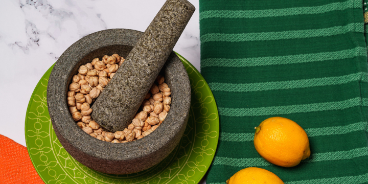 Mortar and pestle with chickpeas and two lemons on marble, counter with orange placemat and green napkin