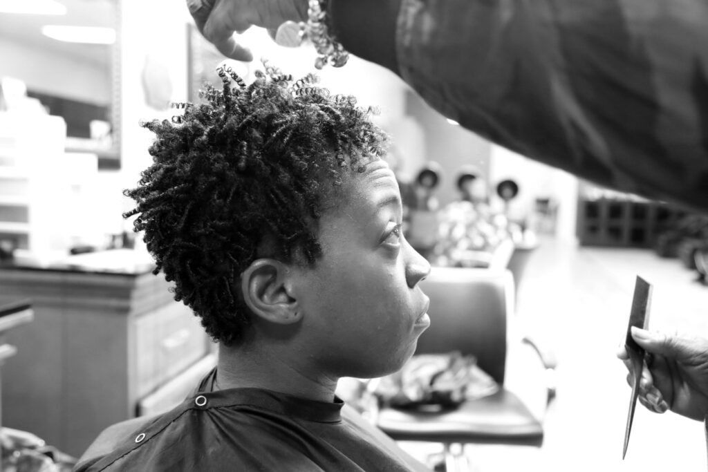Photograph by Zaira Campos. Black and white photo of a young Black person sitting in a barbers chair receiving a haircut.