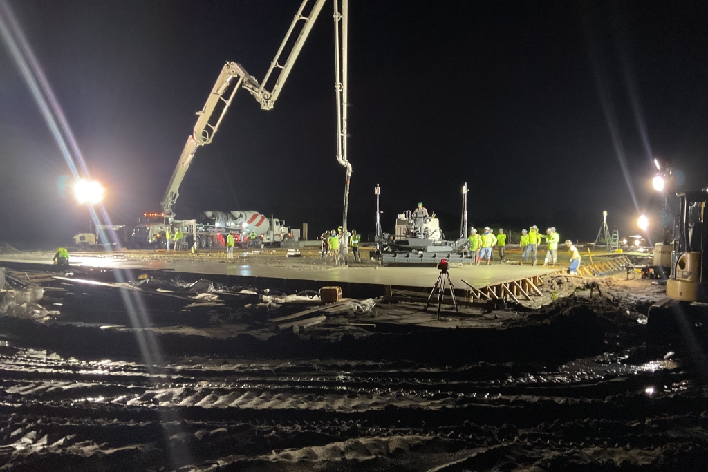Nighttime construction site with workers in high-visibility clothing operating a concrete pump truck.