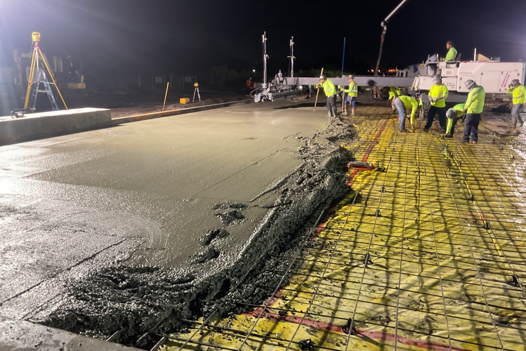 Nighttime construction scene with workers in high-visibility clothing pouring and smoothing concrete under bright lights.
