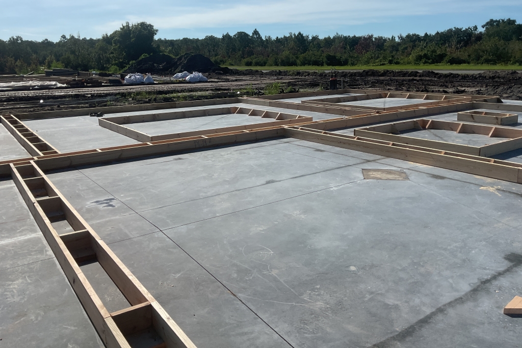 Construction site with a freshly poured concrete foundation sectioned by wooden frames, surrounded by piles of dirt and materials.