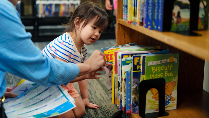A young girl smiles as she chooses a book to read off of a bookshelf.