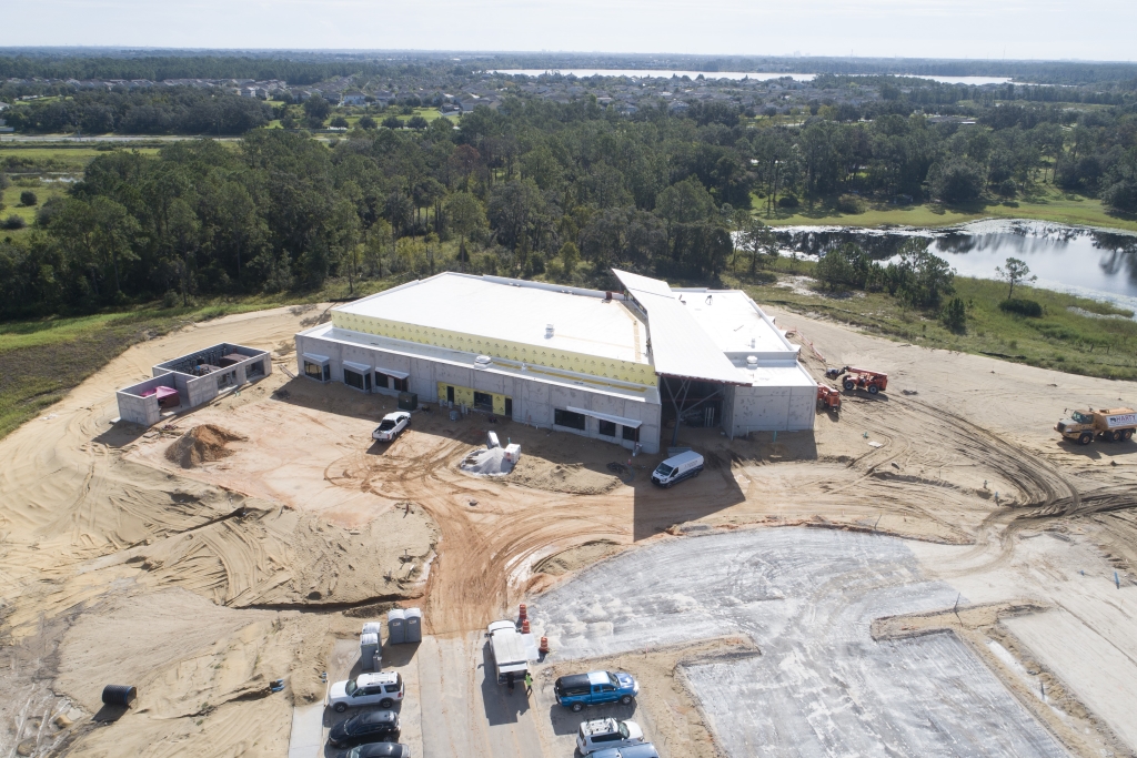 Aerial view of Horizon West construction site with a large white-roofed building, surrounded by dirt, vehicles, and equipment, with trees and a body of water in the background.