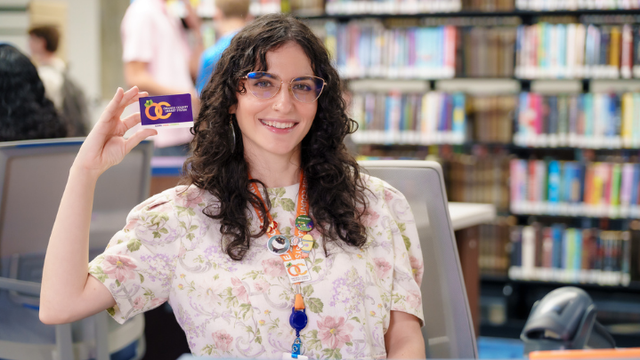 A young woman holds up an Orange County Library System library card while smiling.