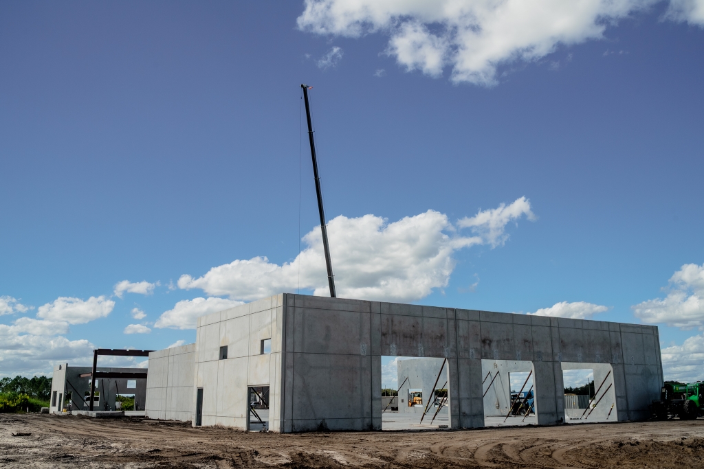 Partially built concrete structure at a construction site with a crane in the background and bare soil in the foreground under a partly cloudy sky.