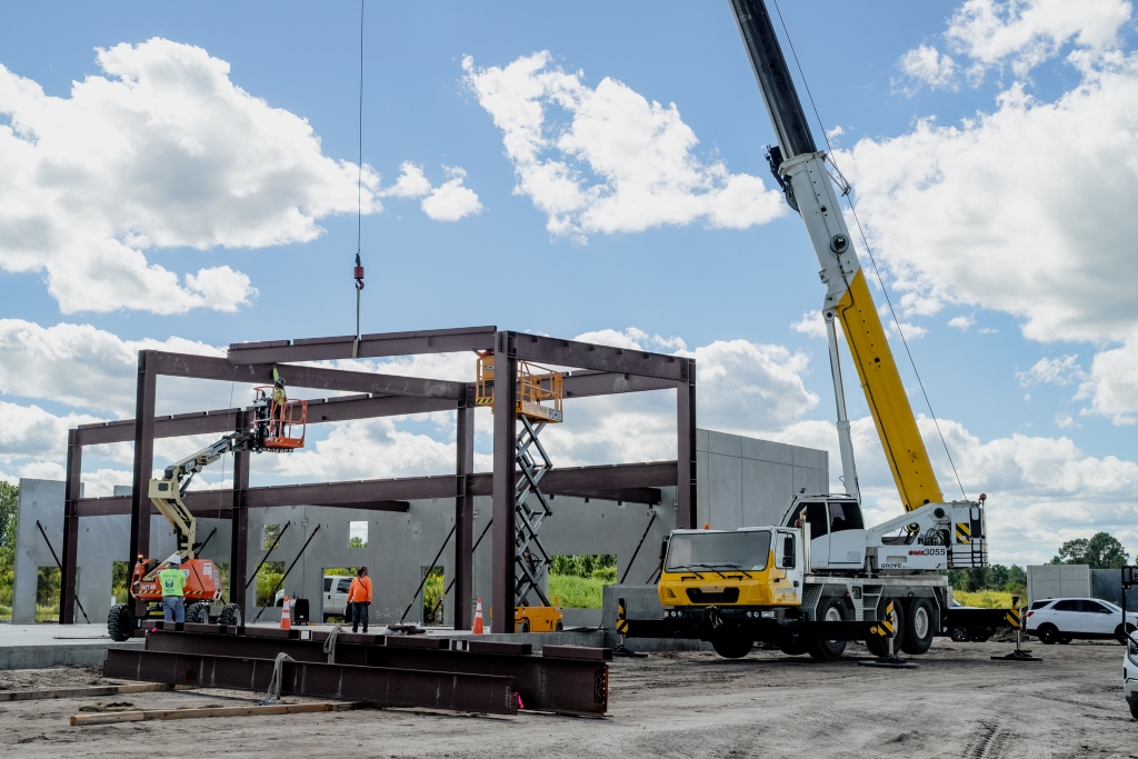 Construction site with workers assembling a steel framework using a crane, cherry picker, and scissor lift under a partly cloudy sky.