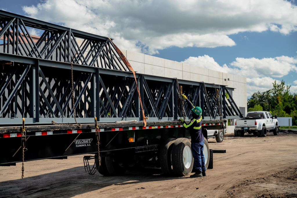 Worker in safety gear securing large black metal trusses onto a flatbed trailer labeled 'Vulcraft' at a construction site, with a white pickup truck and concrete building in the background under a partly cloudy sky.