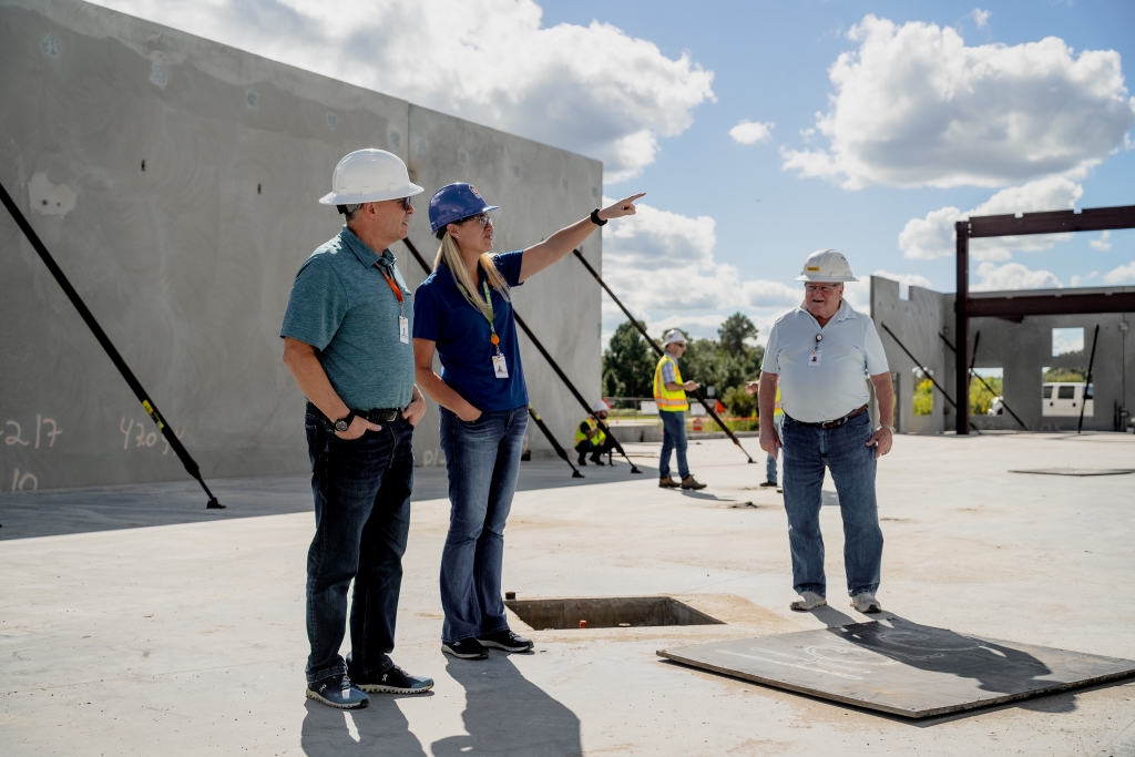 Three library staff and workers in safety helmets and high-visibility vests at a construction site with partially built walls and steel beams, one pointing toward the distance under a partly cloudy sky.