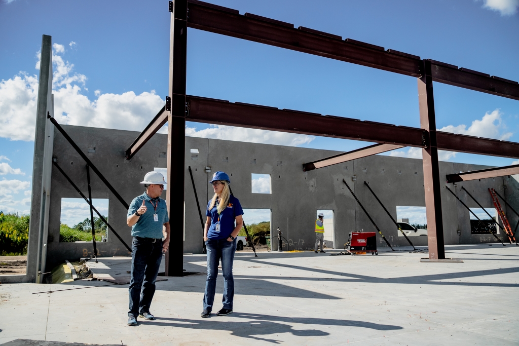 Two library staff and worker in hard hats and safety vests standing on a concrete foundation at a construction site with steel beams and partially built concrete walls under a partly cloudy sky.