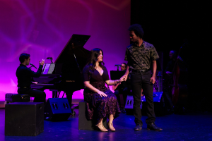 Seated woman and standing man on stage in front of piano player