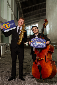 Man holding oversized library card and saxophone stands next to man with Local Wanderer logo sign and stand up bass