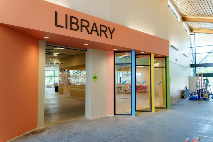 Library entrance with a large orange accent wall displaying the word ‘LIBRARY’ above glass doors framed by colorful panels, opening into an interior space still under construction with equipment and tools nearby.