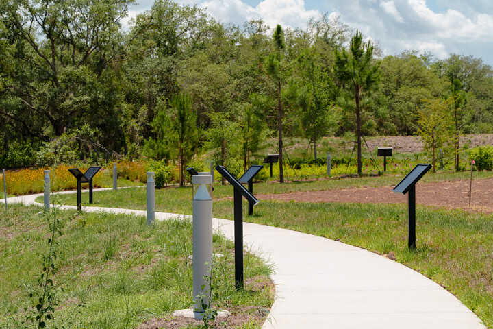 Curved concrete walking path through a landscaped outdoor area with multiple StoryWalk display stands along the route, surrounded by grass, young trees, and natural foliage under a partly cloudy sky.