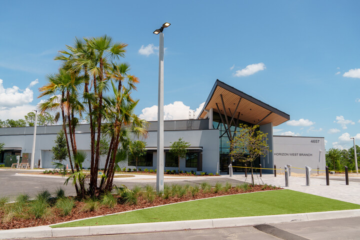 Exterior view of the Horizon West Branch library building with modern architecture, featuring a sloped roof entrance, large windows, landscaped palm trees, and a sidewalk leading to the entrance.