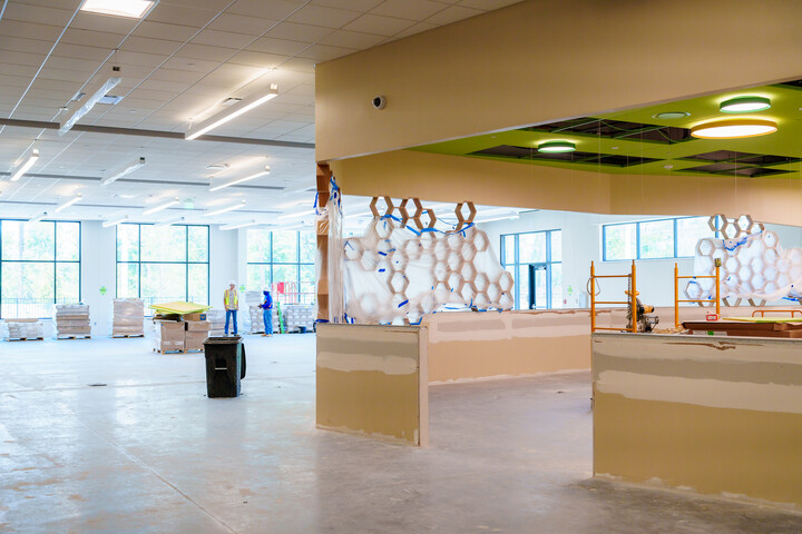 Interior view of a large library space under construction, featuring open floor areas, unfinished service counters, high ceilings with lighting fixtures, large windows, and stacked building materials.
