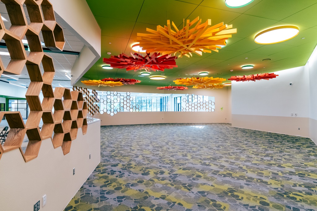 Colorful interior space with patterned carpet, honeycomb wood shelving, and hanging ceiling art in red and yellow.