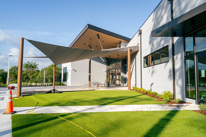 Modern building entrance with a fabric shade canopy under installation, ladders and construction equipment on a landscaped lawn.