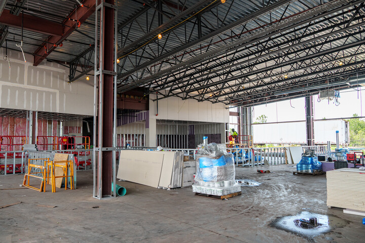 Large open library interior under construction with high steel beam ceiling, construction equipment, stacked materials, scaffolding, and partially built walls across a wide concrete floor.