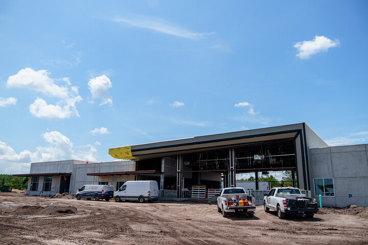 Exterior view of a modern library building under construction with a large open entrance framed by angular architectural elements. Construction vehicles and trucks are parked in front on a dirt lot, with a bright blue sky overhead.