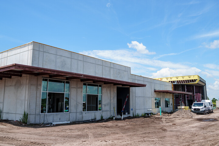 Exterior view of a modern library building under construction with concrete walls, large windows, a covered entrance, construction vehicles, and dirt ground in the foreground under a blue sky.