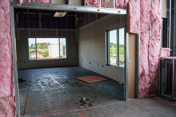 Small unfinished interior room under construction with exposed framing, pink insulation on the walls, concrete floor, and two large windows overlooking an outdoor site.