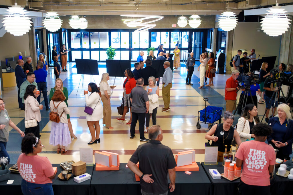 Wide view of a library lobby filled with event attendees, media crews, and display boards, with refreshments set up in the foreground and large decorative light fixtures hanging overhead.