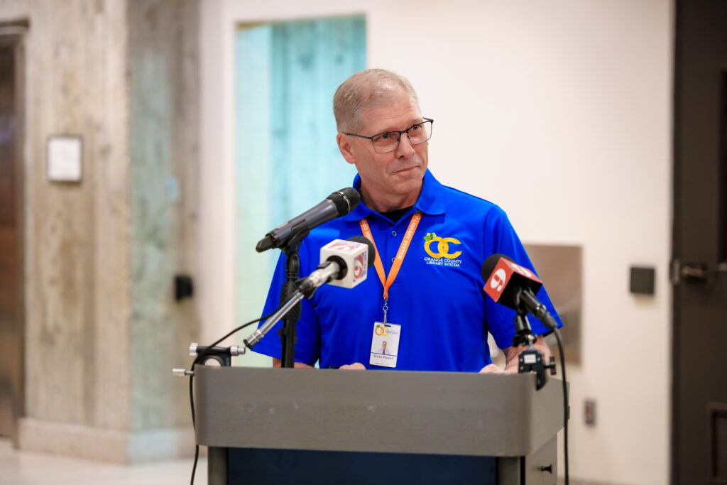 Library Director speaks at a podium during the Orange County Library System first-floor renovation event, with local news microphones positioned in front of the podium.