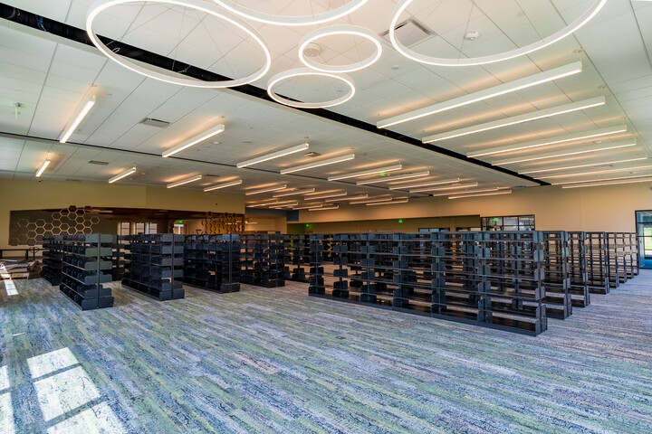 Spacious library collection area with multiple rows of black shelving, patterned carpet, modern circular pendant lights, and large windows providing natural light.