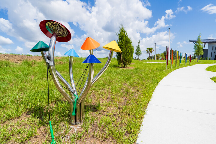 Outdoor learning garden featuring a stainless-steel flower sculpture with colorful cone-shaped elements, set beside a winding concrete path through grassy landscaping.