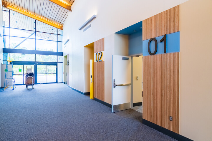 Modern library interior hallway with large glass entrance, high wood-paneled ceiling, and meeting room doors labeled 01 and 02.